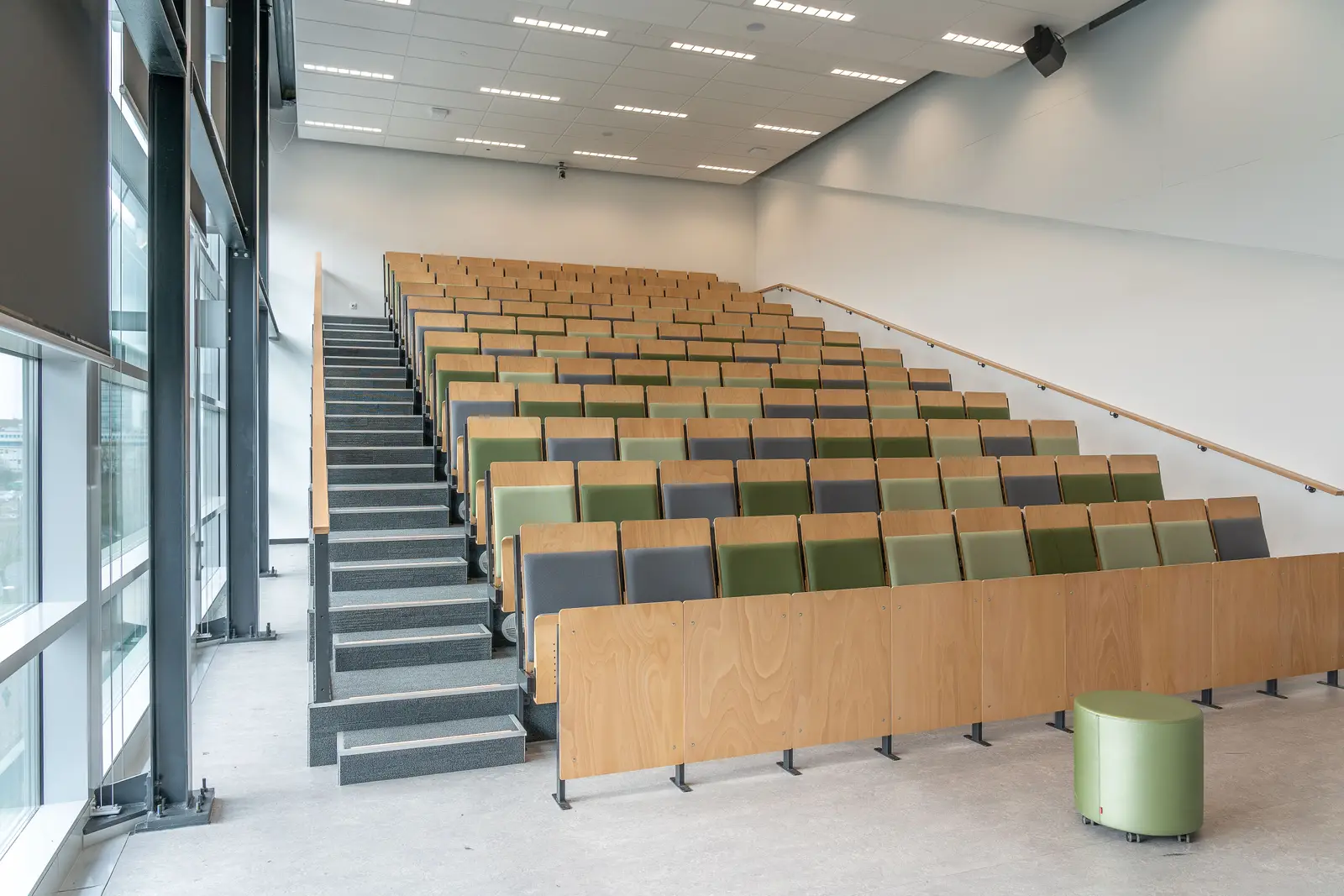 Empty tiered lecture hall with rows of wooden and green seats beside a tall window