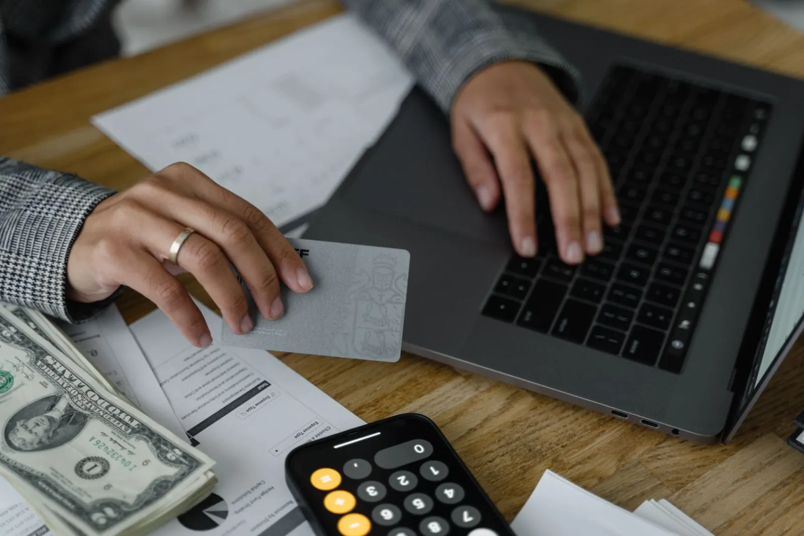 Person holding a credit card while using a laptop with a calculator and cash on the desk