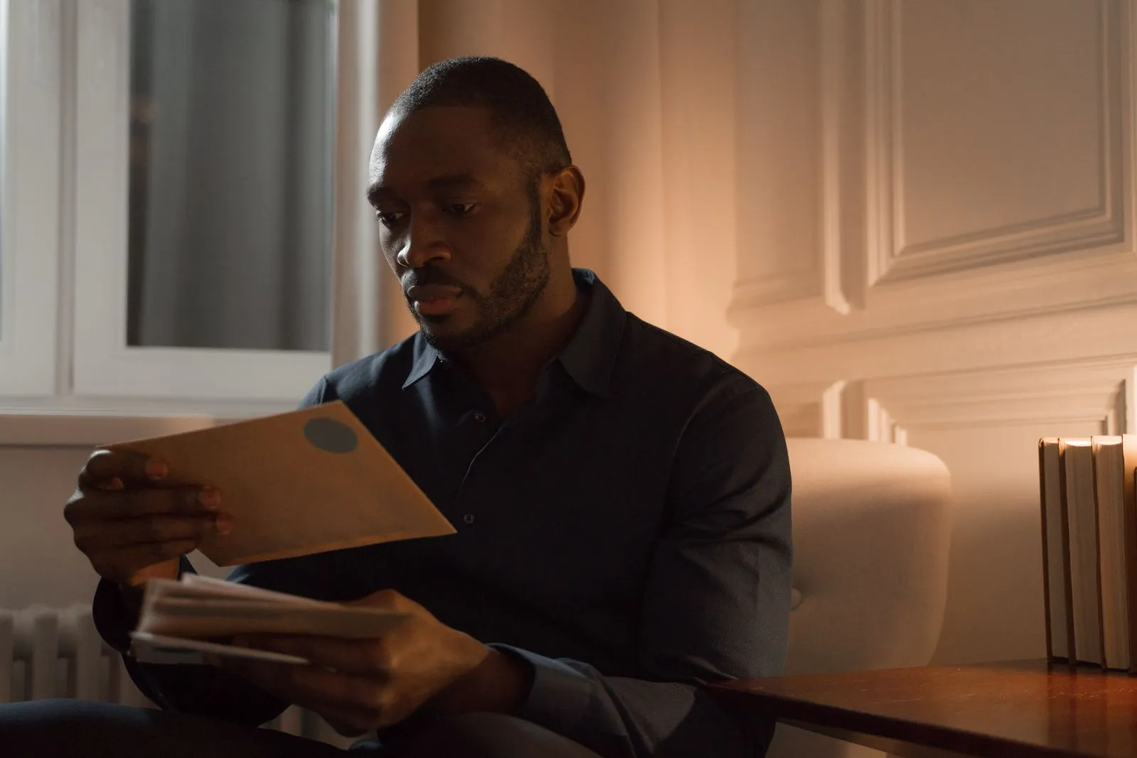 Man in a dark shirt sitting by a window reading a stack of mail