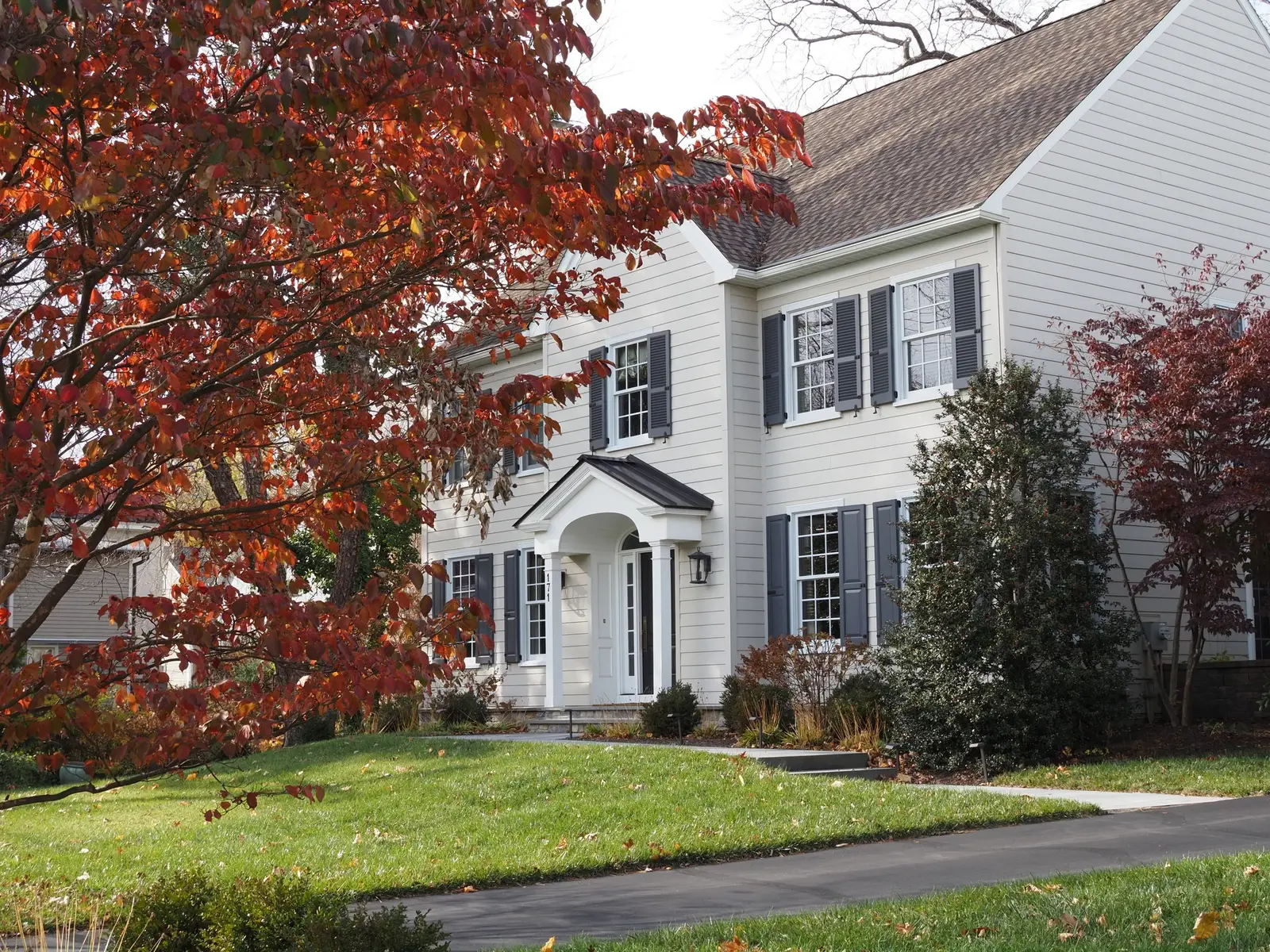 White colonial house with dark shutters behind a red maple in early fall