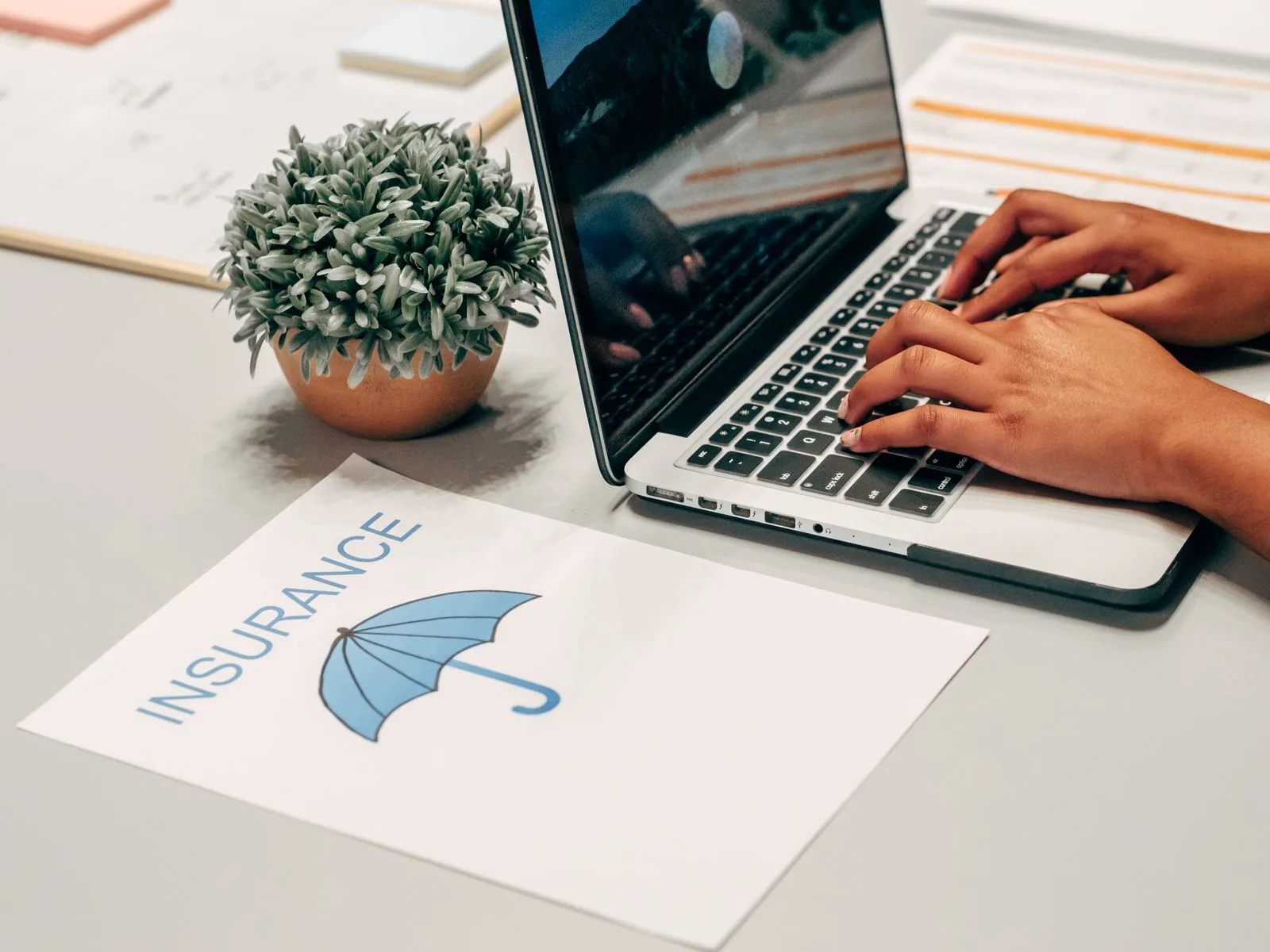 Hands typing on a laptop at a desk beside insurance paperwork and a plant