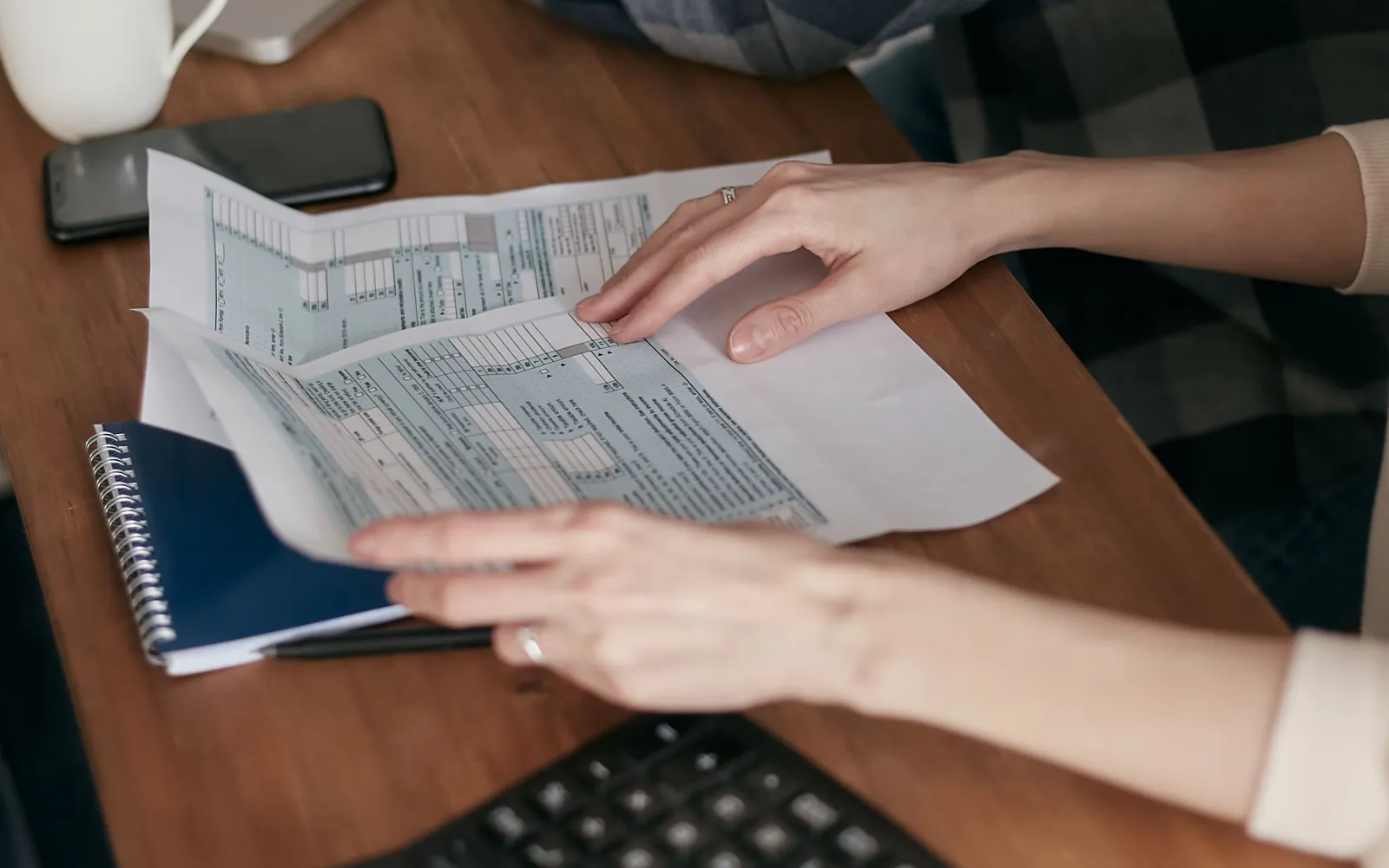 Hands going through tax forms on a wooden desk beside a notebook and calculator