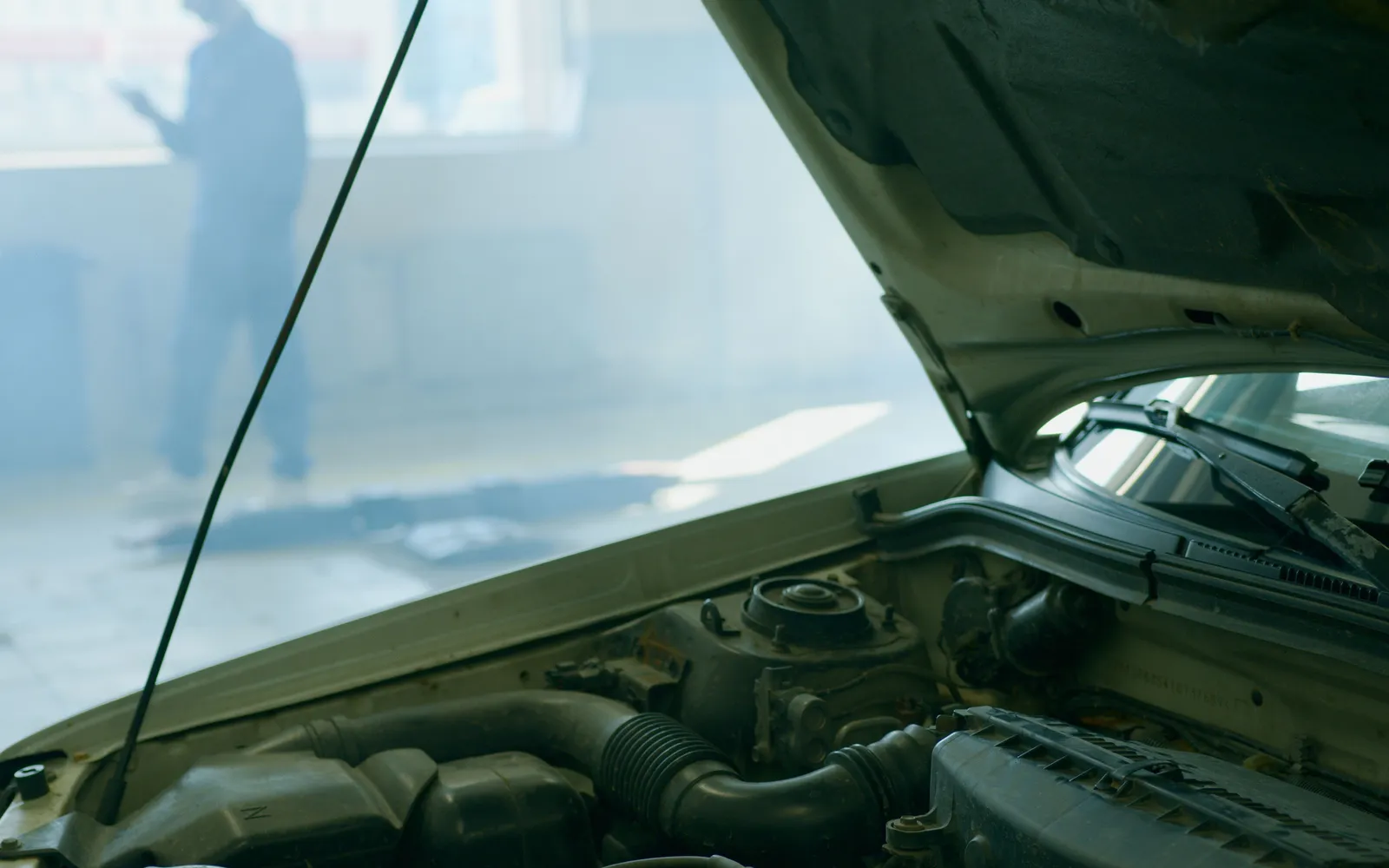 An open car hood in a repair shop with a person holding a device blurred in the background