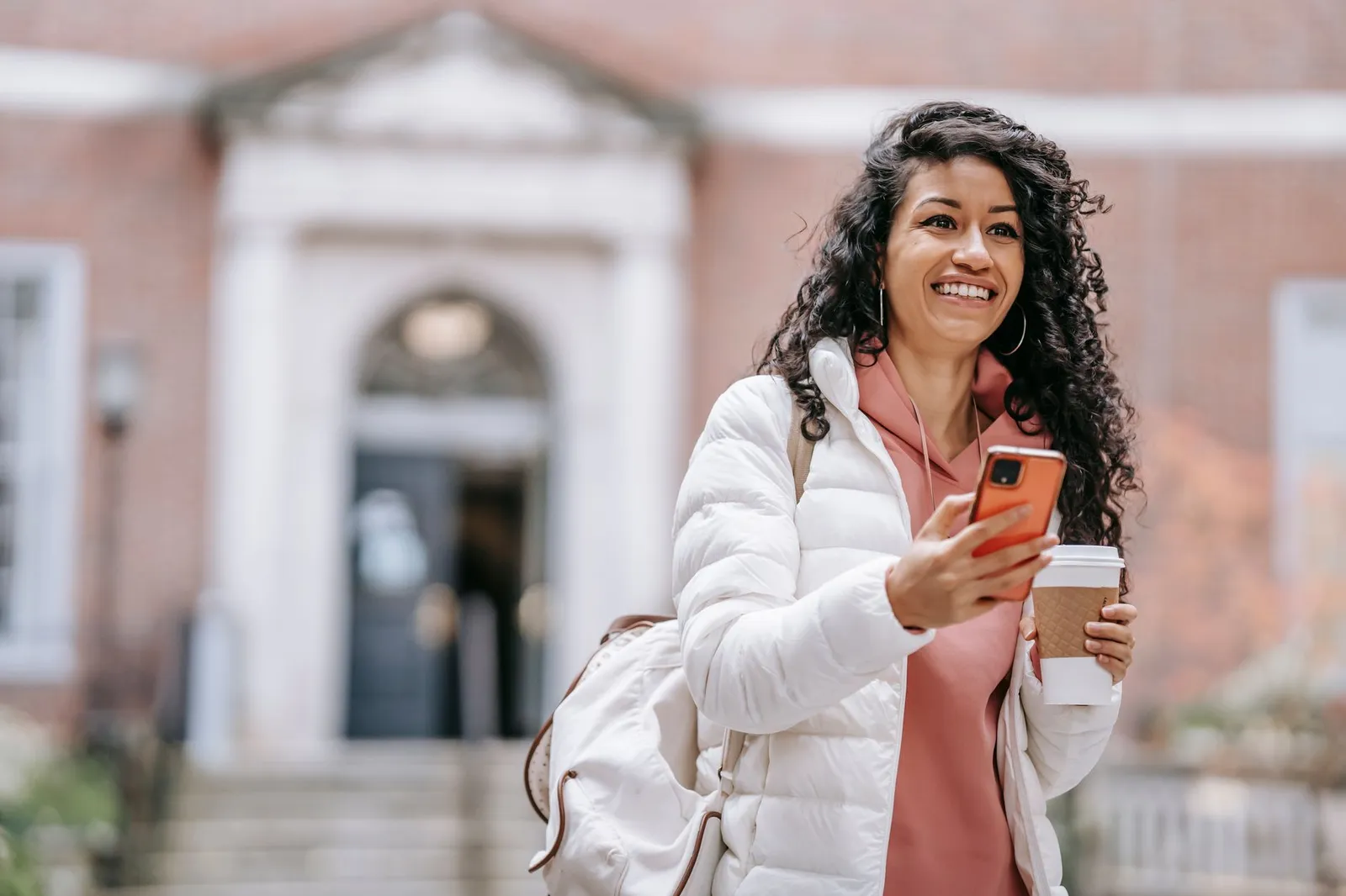 A student holding a phone and a coffee cup outside a brick campus building