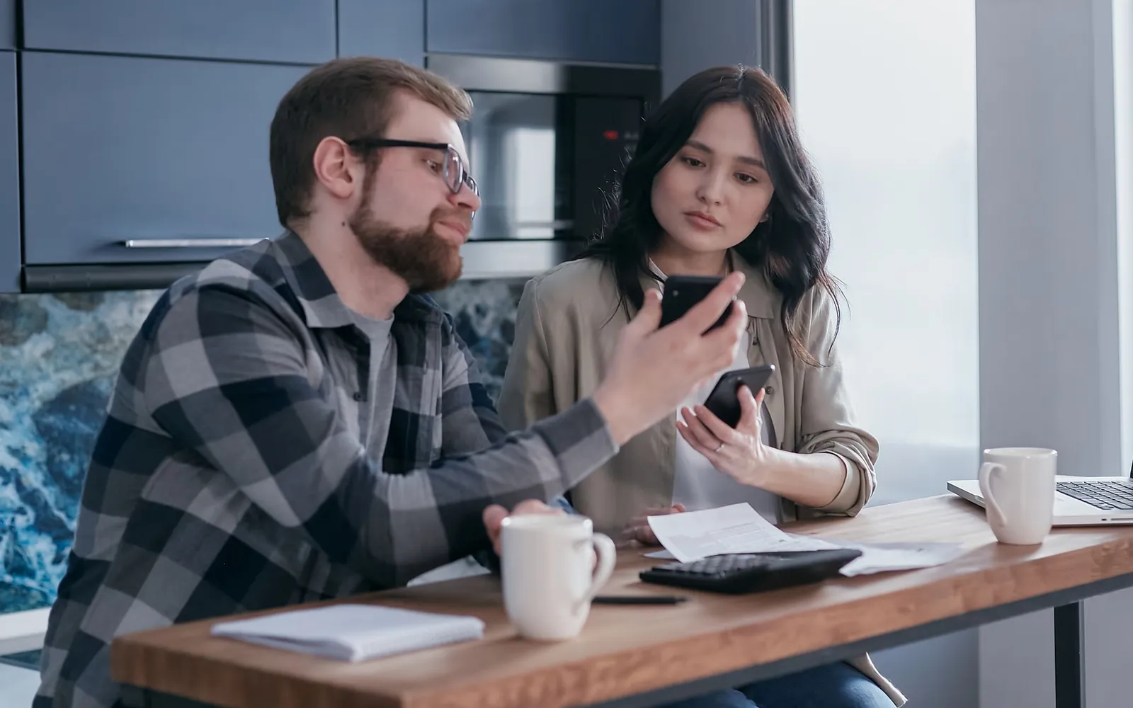 Couple at a kitchen counter frowning at their phones next to a calculator and a printed bill