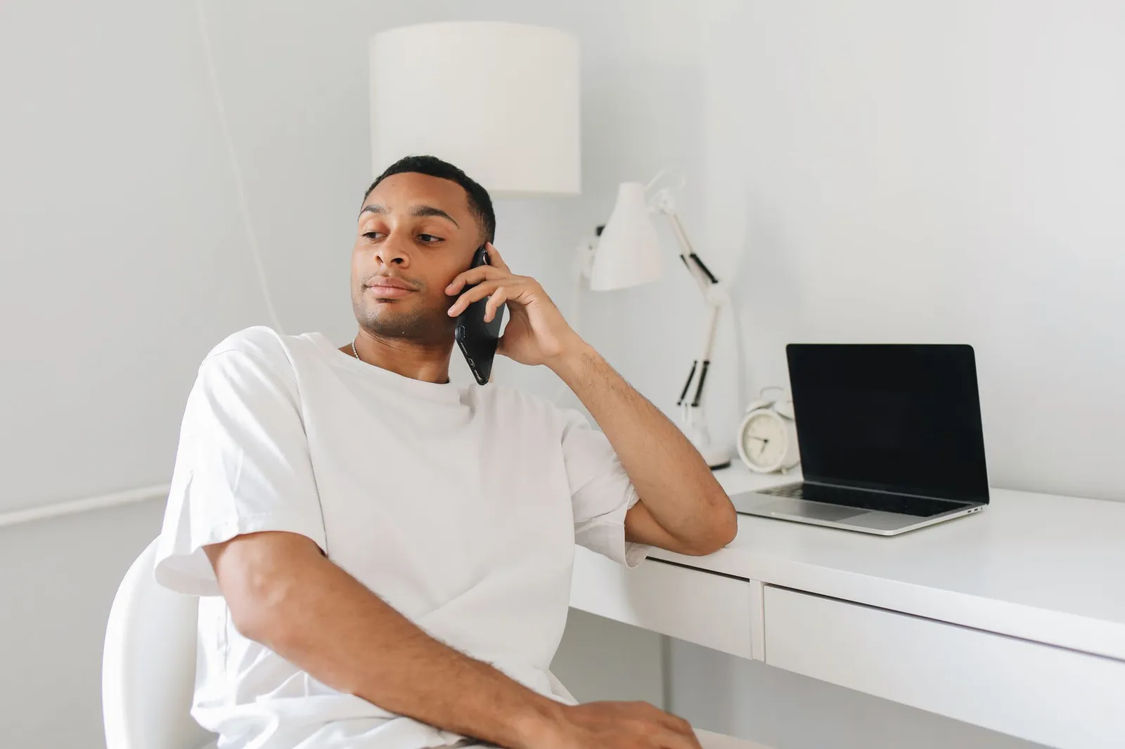 Man sitting at a white desk making a phone call beside a closed laptop
