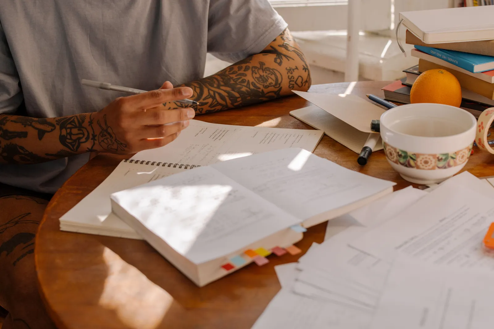 Textbooks, an open notebook and handwritten notes spread across a sunlit wooden desk
