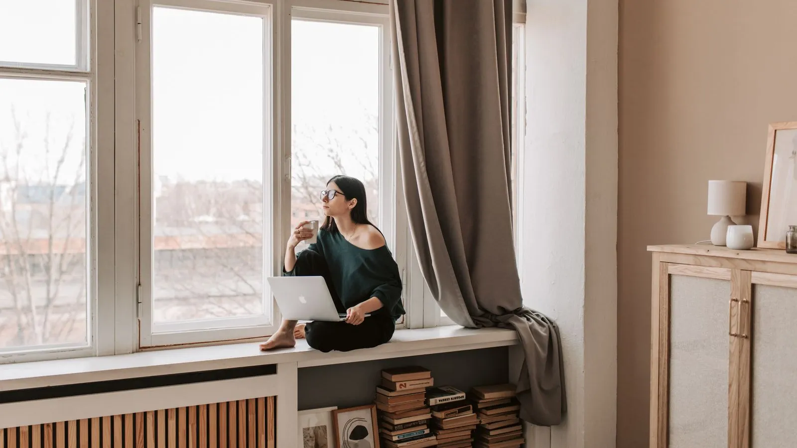 Young woman sitting on a sunlit windowsill with a coffee mug, browsing a laptop