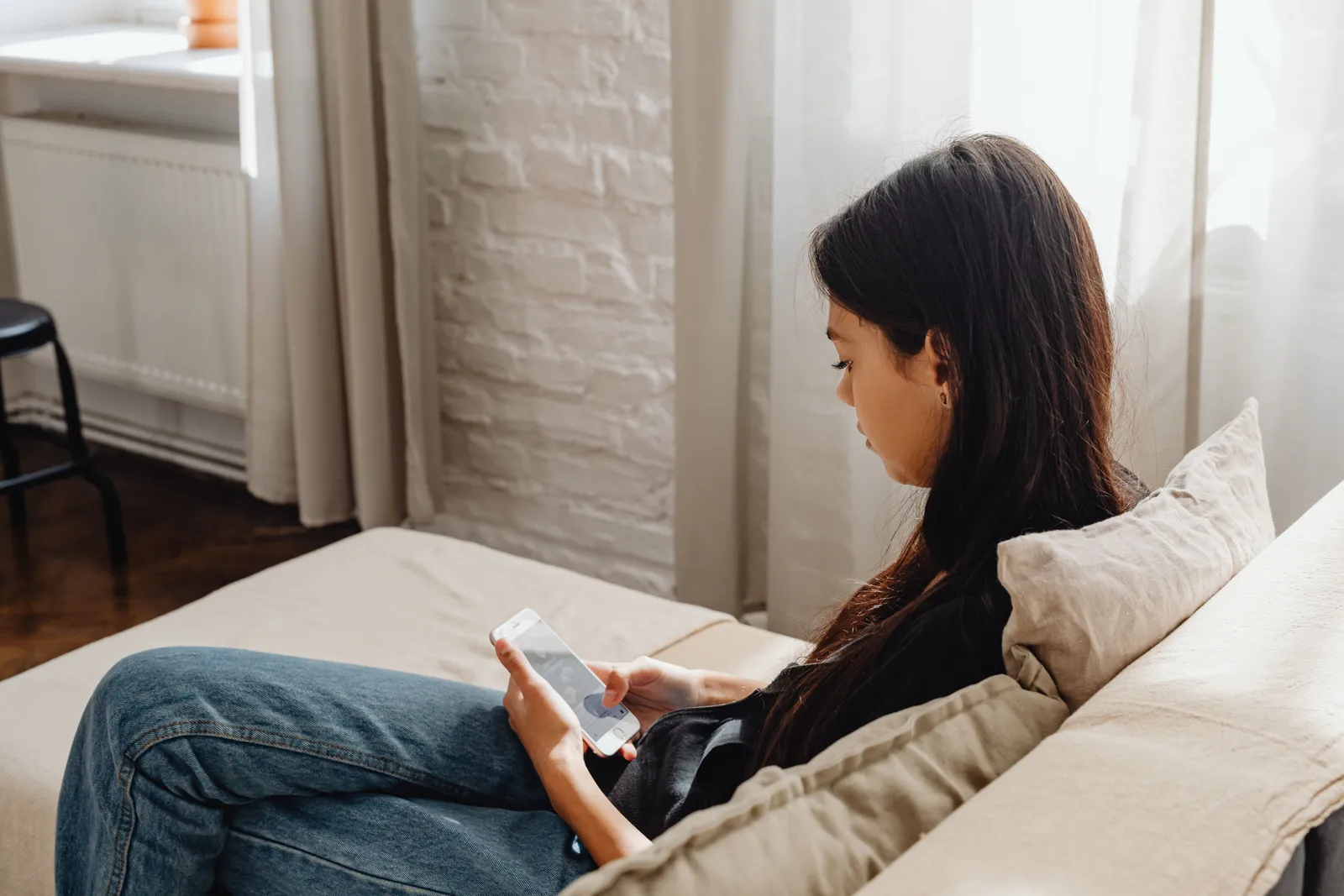 Young woman sitting on a couch in a sunlit apartment, checking her phone