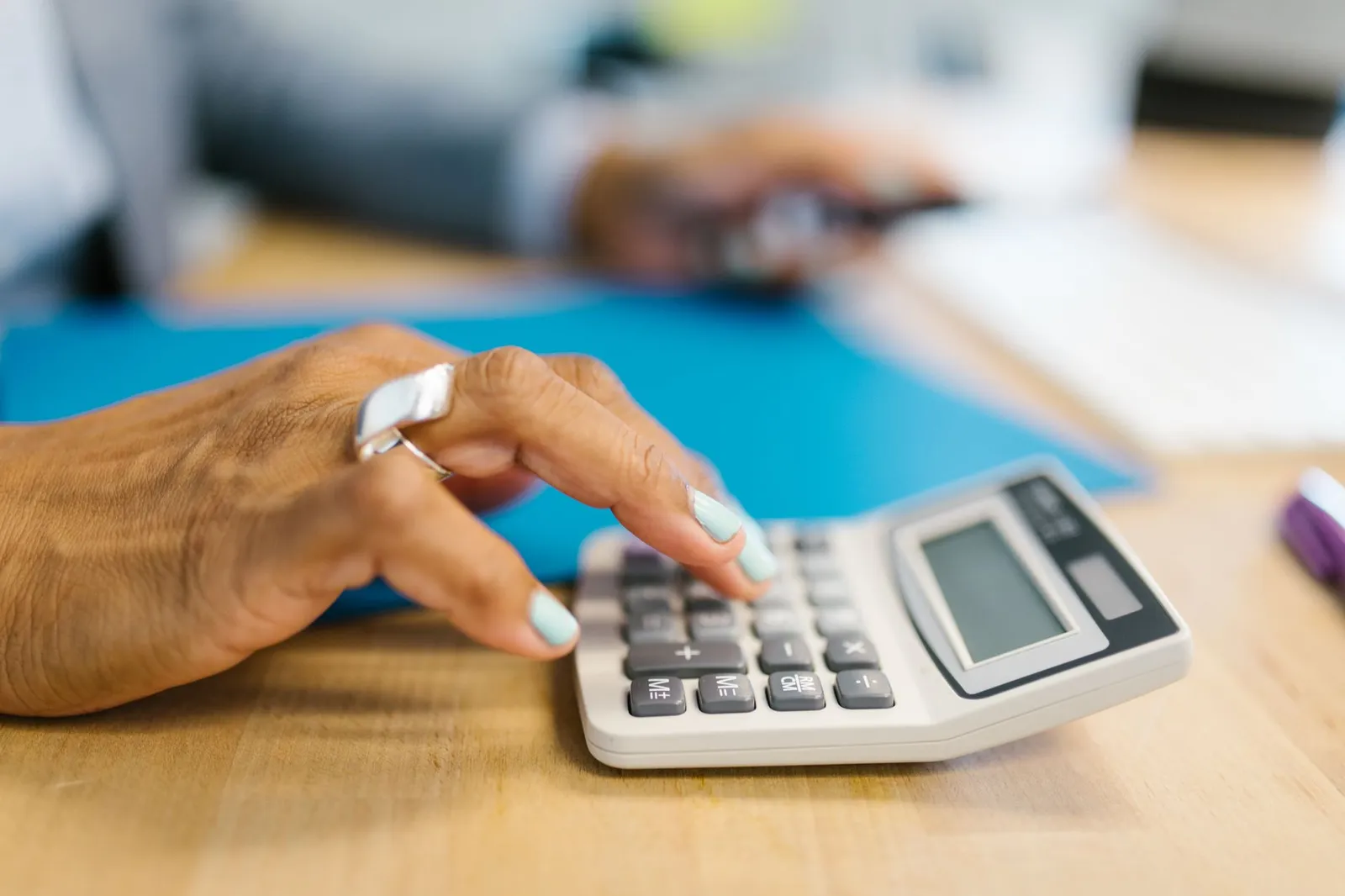 A hand using a desk calculator next to a blue folder on a wooden table
